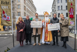 Sechs Erwachsene, einer davon als Nikolaus mit goldenem Umhang und Bischofsstab, stehen auf einer Straße mit großen Schecks in der Hand.