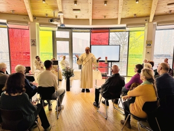 Vor einer Fensterfront mit gelben, roten und klaren Fenstern steht ein Priester. Dahinter ein Bildschirm und ein Altar. Im Vordergrund sind Männer auf Frauen auf Stühlen zu sehen, die mit dem Rücken zur Kamera sitzen.