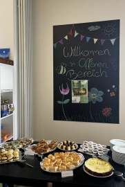 An der Wnad eine Tafel mit Aufschrift "Willkommen", davor ein Buffett mit Kuchen und Snacks.
